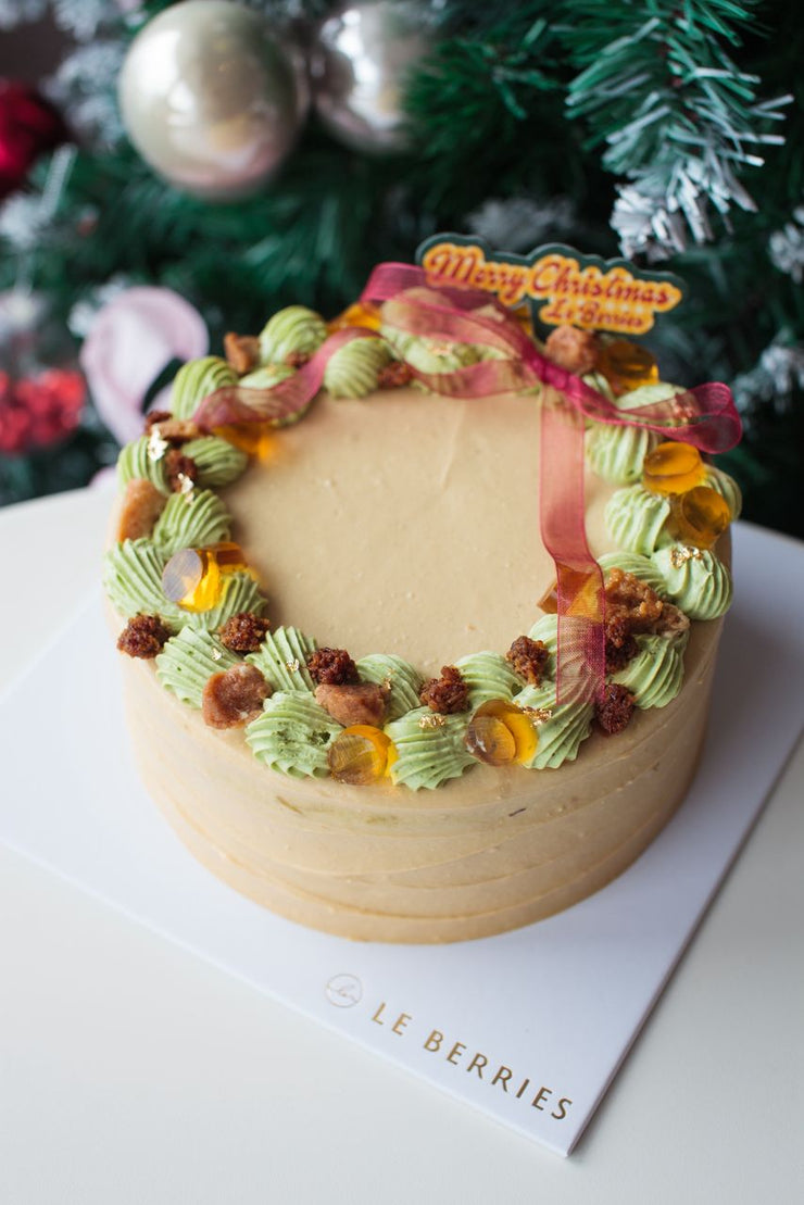 Decorative cake with green icing and festive decorations on a white surface with a Christmas tree in the background.